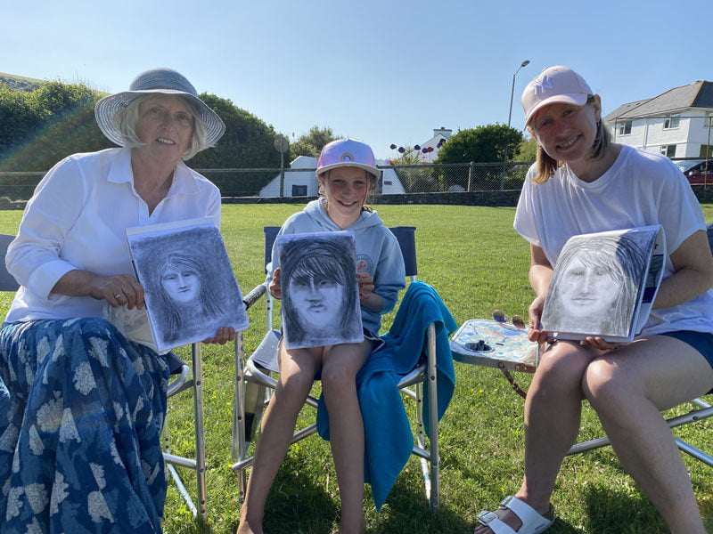 Group art course tuition; three people holding framed pencil portrait artworks outdoors.